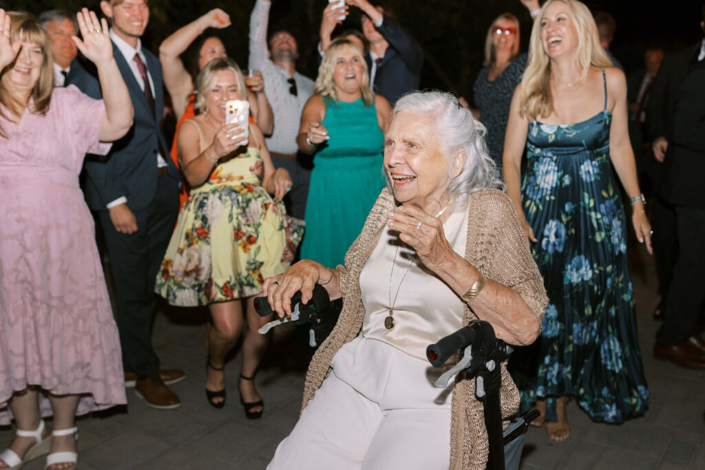 An elderly woman in a wheelchair smiles and laughs while surrounded by a joyful group of people in dresses and suits, celebrating and cheering at an outdoor event. Copyright - Lisa Leanne Photography, California