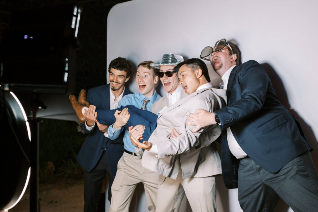 Five men in suits laugh and pose for a photo booth, holding up one of their friends horizontally. Some wear funny props like oversized glasses and a hat, standing in front of a white backdrop with studio lighting. Copyright - Lisa Leanne Photography, California