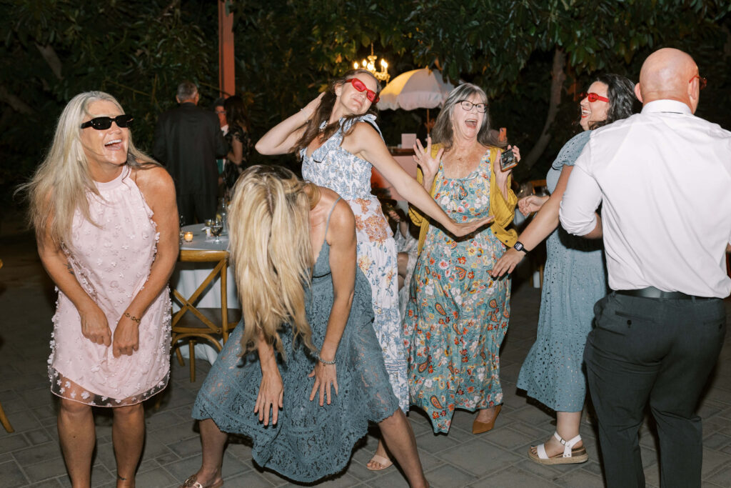 A group of women in colorful dresses and sunglasses dance and laugh together at an outdoor event at night, while a man in a white shirt stands nearby. The mood is lively and joyful. Copyright - Lisa Leanne Photography, California