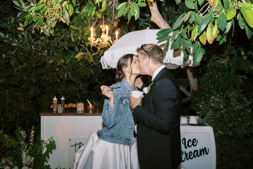 A bride in a denim jacket and a groom in a tuxedo share a kiss in front of an ice cream cart under trees at night, with a chandelier and umbrella above them. The bride holds a cup of ice cream and a spoon. Copyright - Lisa Leanne Photography, California