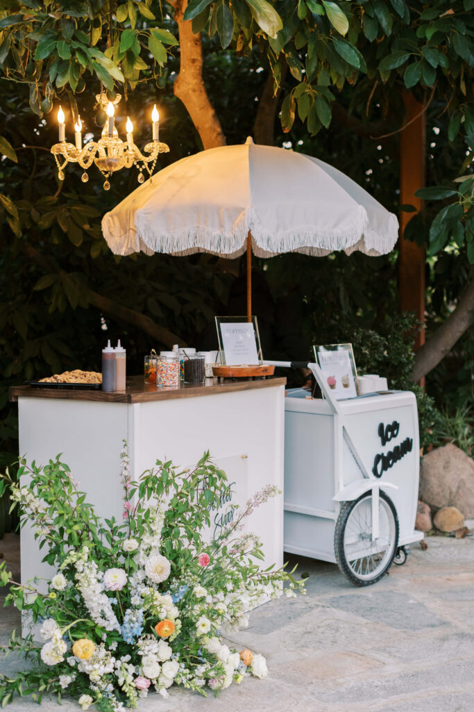 A charming outdoor ice cream cart with a white umbrella and chandelier, surrounded by lush greenery and a colorful floral arrangement. Jars of toppings and an 