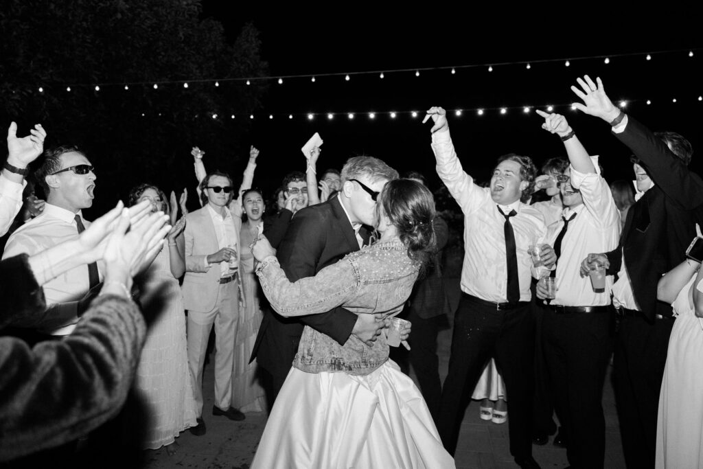 A couple kisses on a dance floor at night, surrounded by cheering and celebrating guests. String lights hang overhead, and many people have their arms raised in excitement. The scene is joyful and energetic. Copyright - Lisa Leanne Photography, California