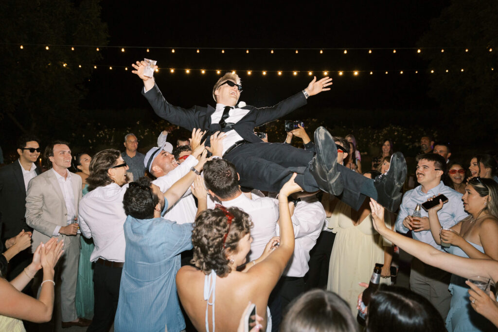 A group of partygoers lift a man in a suit and sunglasses into the air at a nighttime celebration under string lights; people around him are cheering and clapping. Copyright - Lisa Leanne Photography, California