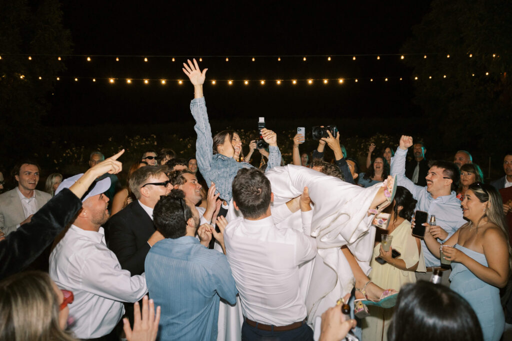 A joyful crowd lifts a smiling woman in a white dress during a nighttime celebration, with string lights glowing overhead and people cheering and taking photos. Copyright - Lisa Leanne Photography, California