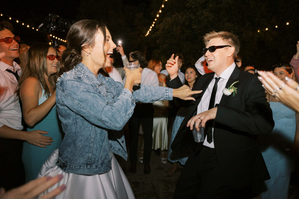 A joyful bride in a denim jacket and a man in a black suit and sunglasses dance together at a lively outdoor celebration, surrounded by smiling guests under string lights. Copyright - Lisa Leanne Photography, California