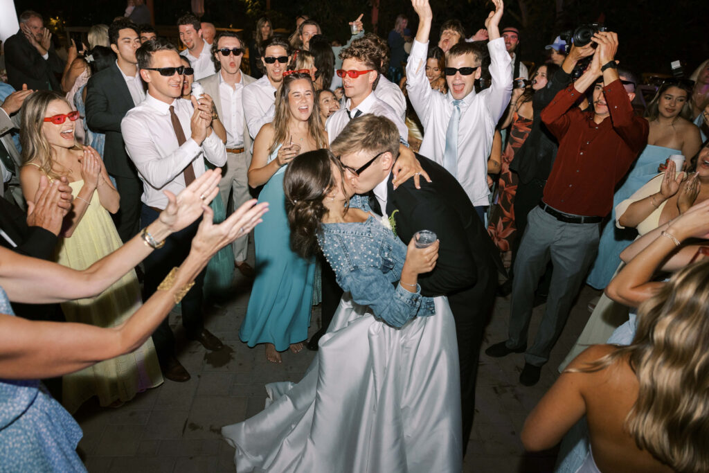 A couple shares a kiss on the dance floor surrounded by cheering guests; many wear sunglasses and colorful attire, creating a lively and celebratory wedding atmosphere. Copyright - Lisa Leanne Photography, California