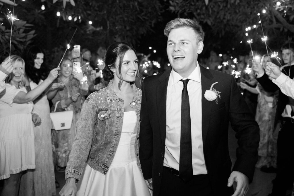 A smiling bride in a dress and denim jacket walks hand-in-hand with a groom in a suit. They are surrounded by cheering guests holding sparklers at an outdoor nighttime celebration. Copyright - Lisa Leanne Photography, California