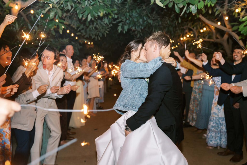 A bride and groom kiss under string lights as guests cheer and hold sparklers around them at an outdoor evening wedding celebration. Copyright - Lisa Leanne Photography, California