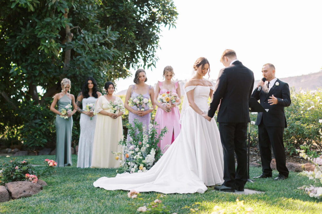 A bride and groom stand holding hands during an outdoor wedding ceremony. A man officiates while six bridesmaids in pastel dresses stand nearby, each holding bouquets. The scene is lush with greenery and flowers. Copyright - Lisa Leanne Photography, California