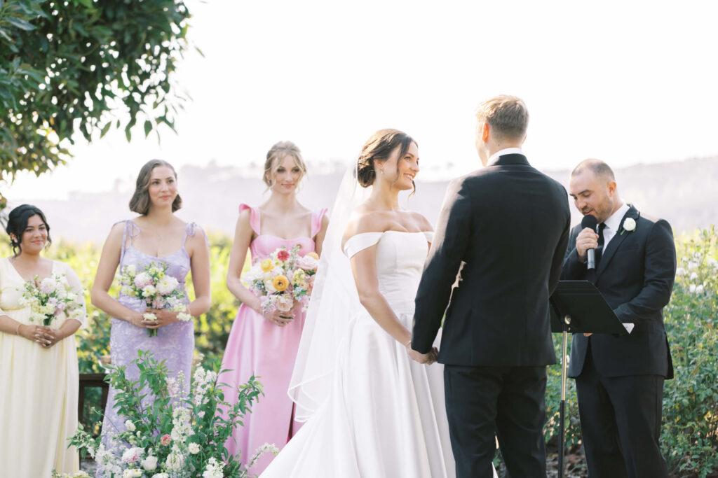 A bride and groom stand holding hands during an outdoor wedding ceremony, with a man officiating. Bridesmaids in pastel dresses holding bouquets stand to the side, surrounded by greenery and flowers. Copyright - Lisa Leanne Photography, California