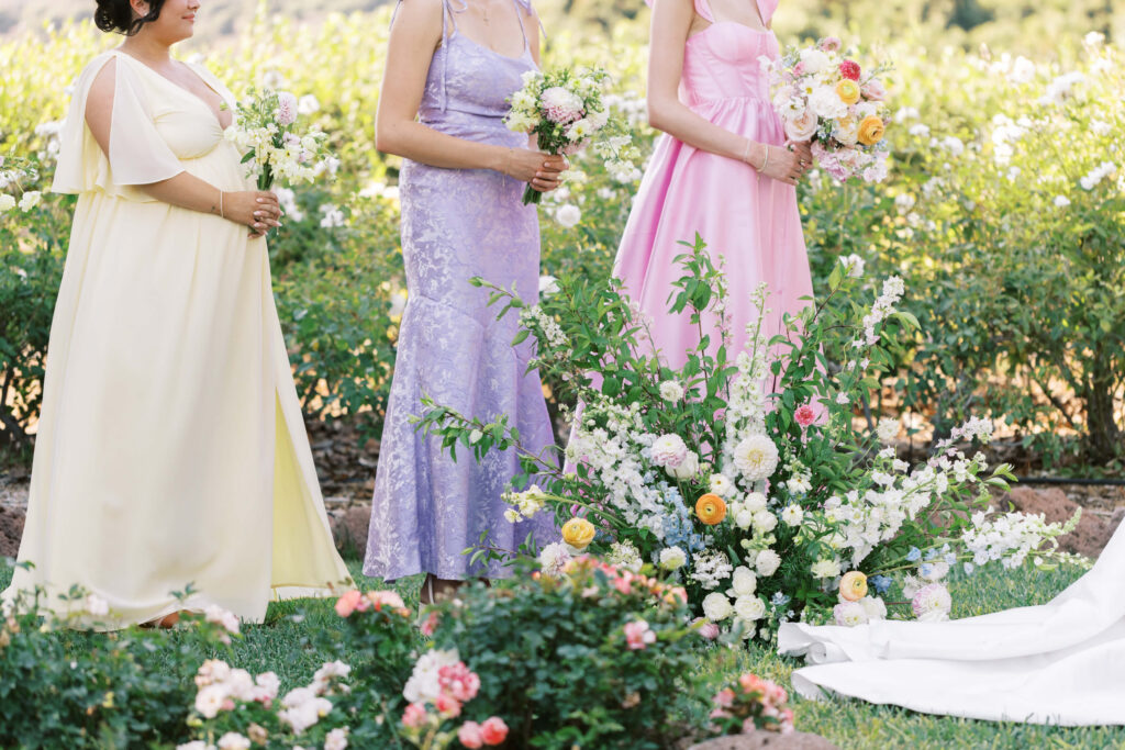 Three women in pastel dresses—yellow, lavender, and pink—stand outdoors holding bouquets. They are surrounded by lush greenery and flowers, with additional floral arrangements at their feet. Their faces are not visible. Copyright - Lisa Leanne Photography, California