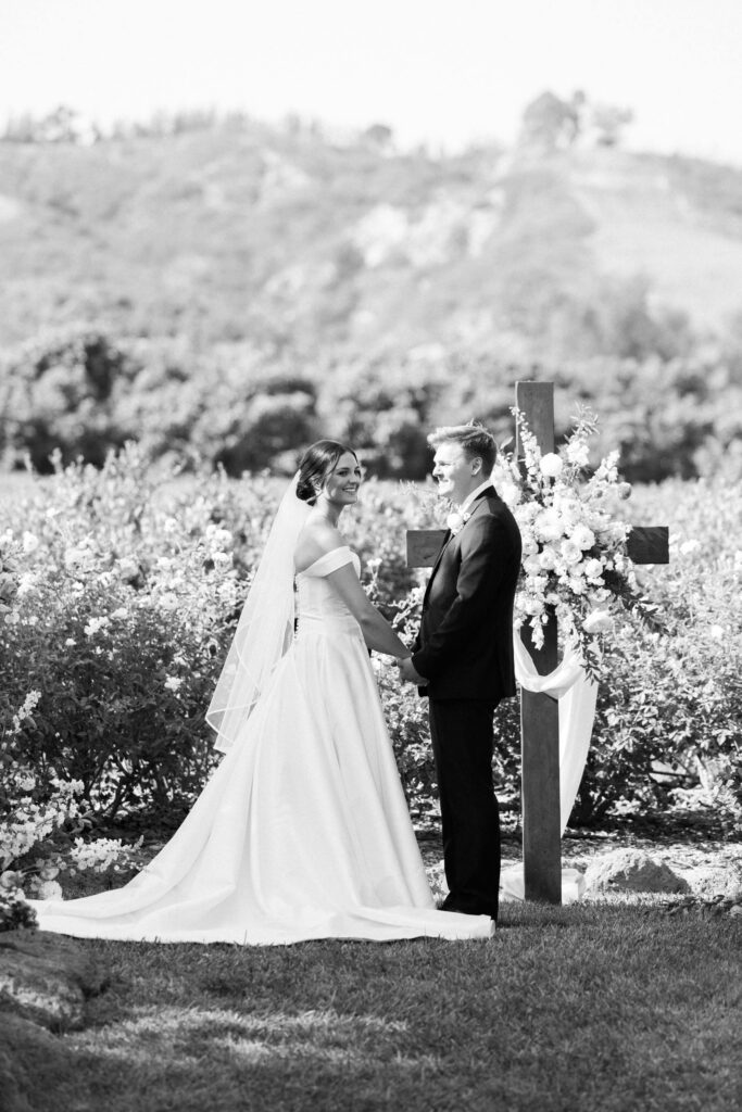 A bride and groom stand holding hands outdoors in front of a large floral cross, smiling at each other. The bride wears an off-shoulder gown and veil, and the groom is in a suit. Trees and hills are in the background. Copyright - Lisa Leanne Photography, California