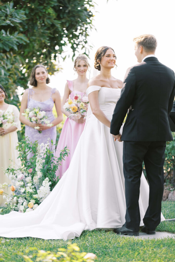 A bride in a white gown and a groom in a black suit stand holding hands outdoors during a wedding ceremony, with bridesmaids in pastel dresses holding bouquets in the background. Copyright - Lisa Leanne Photography, California