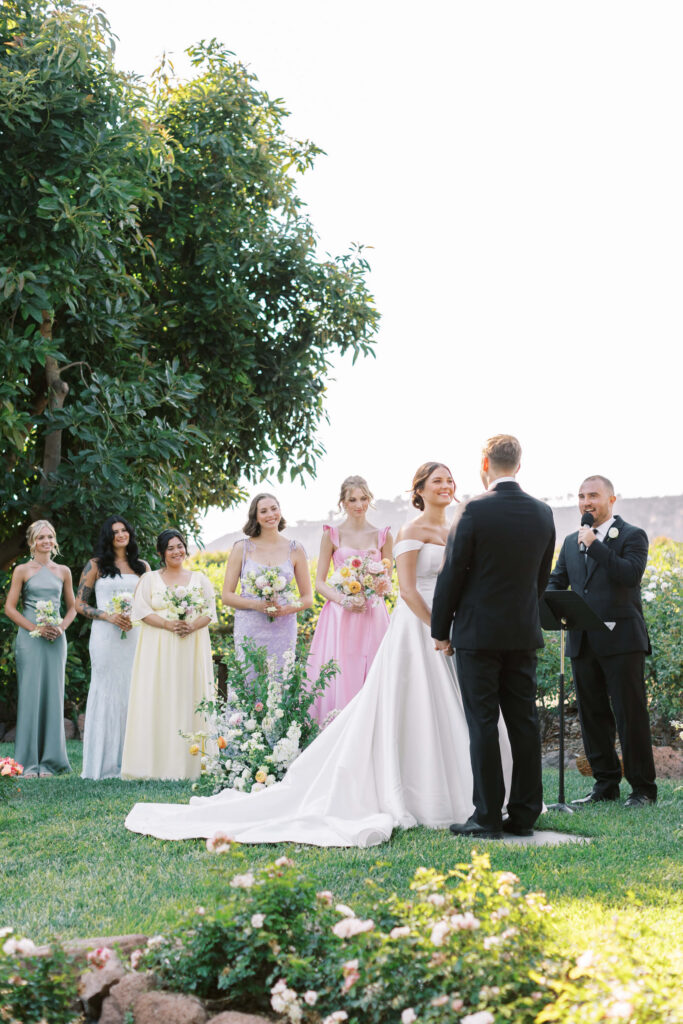A bride and groom stand facing each other during an outdoor wedding ceremony, with bridesmaids in pastel dresses holding bouquets and an officiant speaking beside them. Greenery and flowers surround the scene. Copyright - Lisa Leanne Photography, California