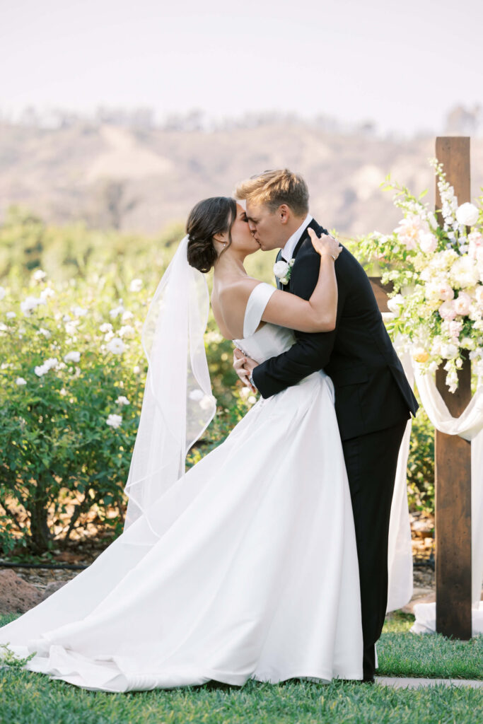 A bride and groom share a kiss outdoors during their wedding ceremony, surrounded by greenery and white flowers. The bride wears an off-shoulder white gown and veil; the groom is dressed in a black suit and bow tie. Copyright - Lisa Leanne Photography, California