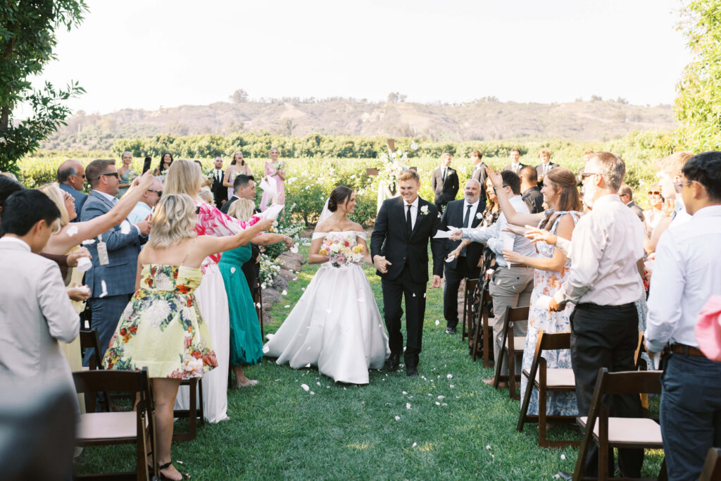 A bride and groom walk down an outdoor aisle, smiling, as guests stand on either side, tossing flower petals and celebrating on a sunny day. Green hills and fields are visible in the background. Copyright - Lisa Leanne Photography, California
