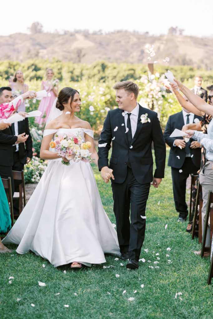A bride and groom walk down the aisle outdoors, smiling at each other, as guests toss flower petals. The bride wears a white off-shoulder gown and holds a bouquet; the groom wears a black suit. Greenery and hills are visible in the background. Copyright - Lisa Leanne Photography, California