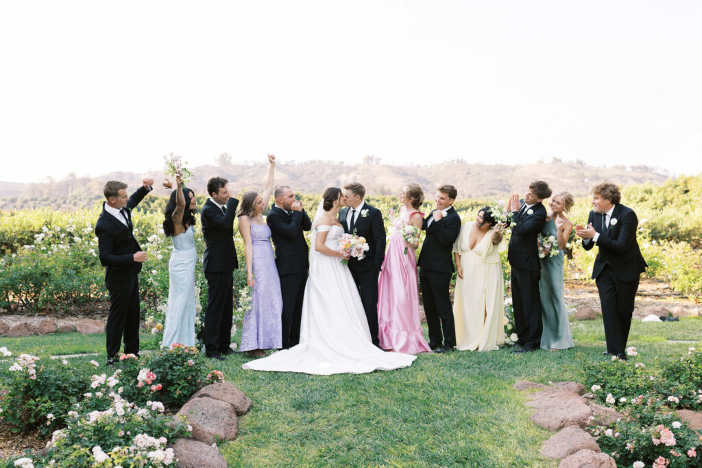 A bride and groom kiss outdoors, surrounded by their wedding party in formal attire, with bridesmaids in pastel dresses and groomsmen in black suits, celebrating amid gardens and distant hills. Copyright - Lisa Leanne Photography, California