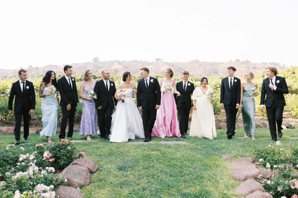 A wedding party walks outdoors on a grassy lawn, with bridesmaids in pastel dresses and groomsmen in black suits. The bride and groom are centered, and everyone appears joyful, with greenery and hills in the background. Copyright - Lisa Leanne Photography, California
