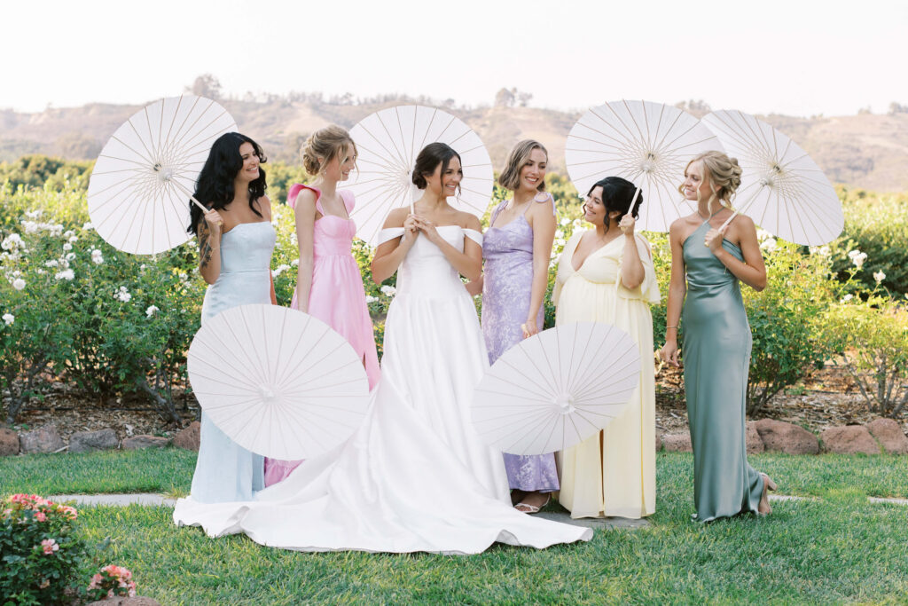 Six women in formal dresses stand outdoors holding white parasols. The woman in the center wears a white wedding gown, surrounded by bridesmaids in pastel dresses, all smiling and talking in a garden setting. Copyright - Lisa Leanne Photography, California