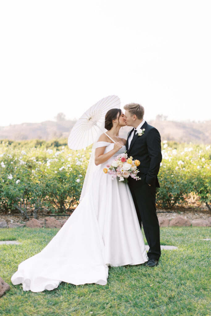 A bride in a white dress holding a parasol and a bouquet kisses a groom in a black suit outdoors, with greenery and flowers in the background. Copyright - Lisa Leanne Photography, California