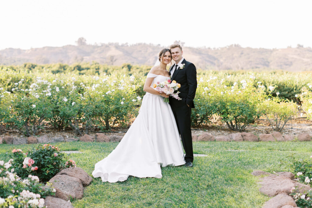 A bride in a white off-shoulder gown and groom in a black suit stand together outdoors, smiling and holding a bouquet, with green bushes and hills in the background. Copyright - Lisa Leanne Photography, California