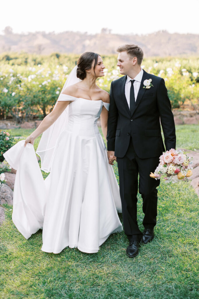 A bride in a white off-shoulder gown and veil holds hands with a groom in a black suit and tie. They are smiling at each other while walking on grass in a garden, with flowers and hills in the background. Copyright - Lisa Leanne Photography, California