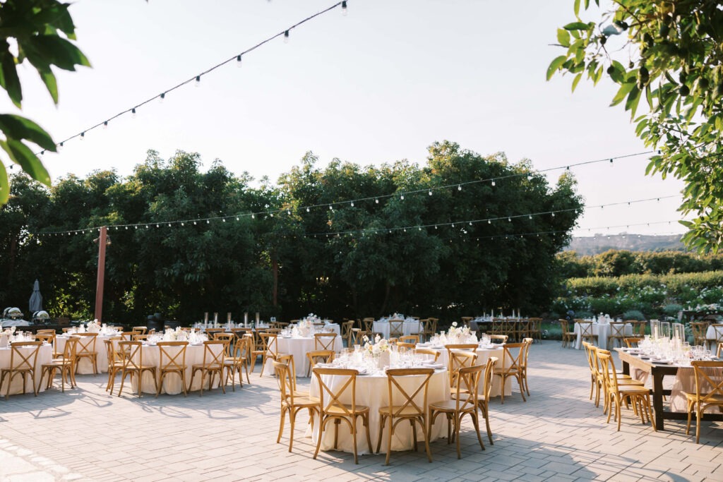 Outdoor event setup with round tables and wooden chairs arranged on a paved patio, decorated with white tablecloths and string lights overhead, surrounded by green trees under clear skies. Copyright - Lisa Leanne Photography, California