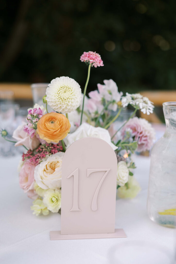 A white table centerpiece with pastel flowers and greenery, next to a table number sign reading 