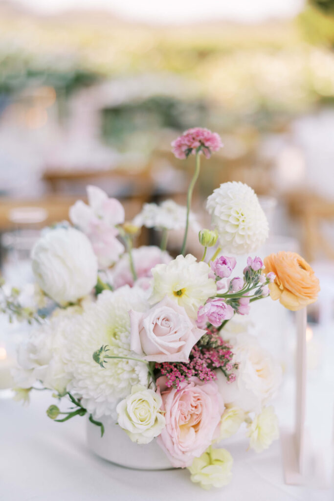 A pastel-colored floral arrangement including white dahlias, blush roses, pale pink flowers, and a single orange ranunculus sits in a white vase on a table, with a softly blurred outdoor background. Copyright - Lisa Leanne Photography, California