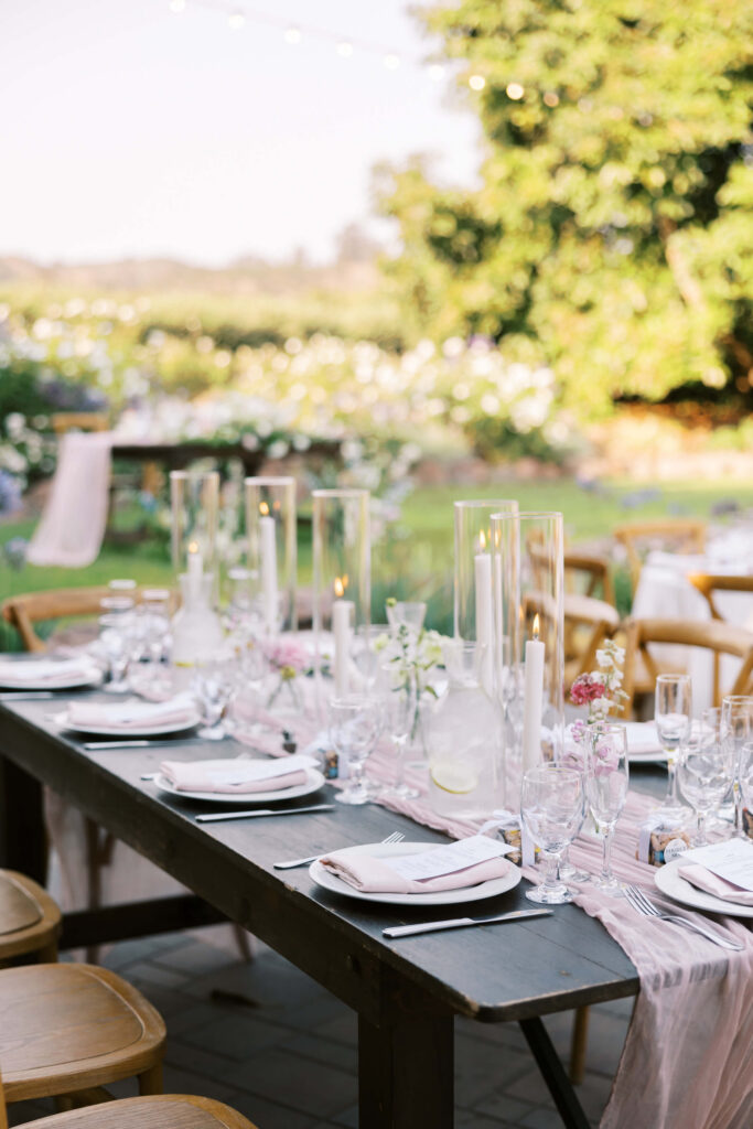 An elegant outdoor table set for a formal event, featuring white plates, crystal glasses, folded napkins, and tall glass candle holders with white candles, surrounded by greenery and flowers in a garden setting. Copyright - Lisa Leanne Photography, California
