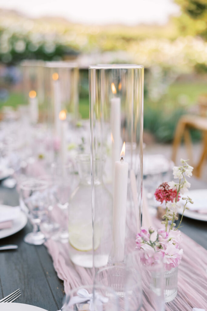 A close-up of an elegant outdoor table setting with tall glass candle holders containing lit white candles, glassware, a pitcher of water with lemon, and small vases of pink and white flowers. Copyright - Lisa Leanne Photography, California