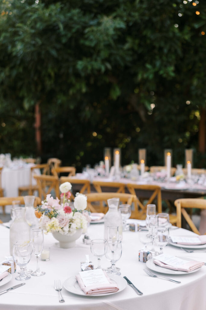 Elegant outdoor table setting with white tablecloths, floral centerpieces, glassware, plates, and folded napkins. Wooden chairs and candles create a warm, inviting atmosphere, with greenery in the background. Copyright - Lisa Leanne Photography, California