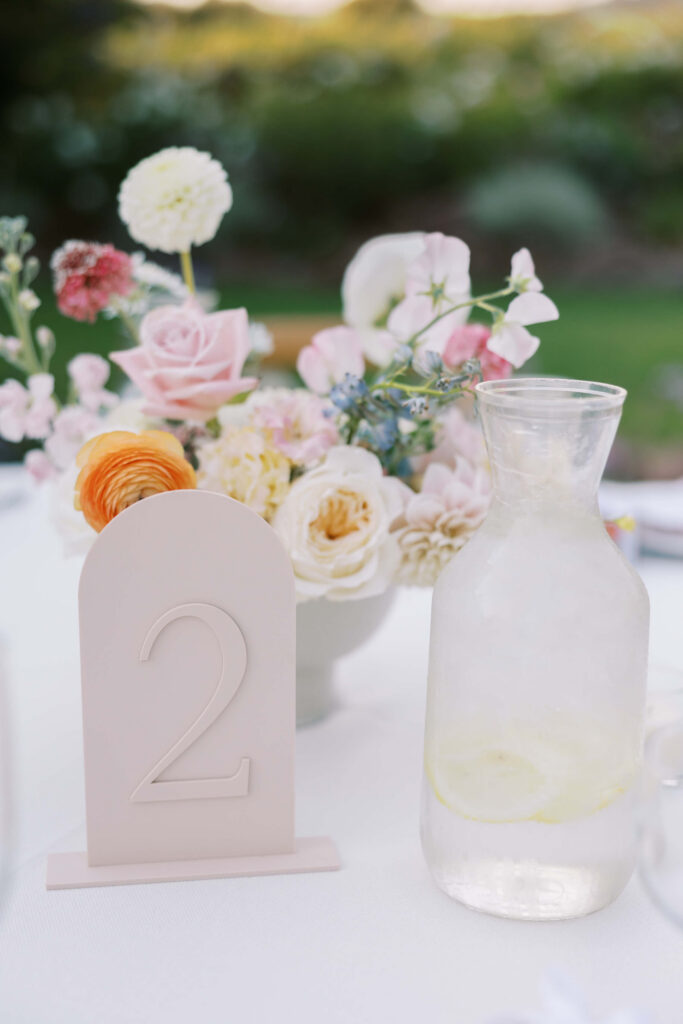 A table centerpiece with a vase of pastel flowers, a pale pink table number 2 sign, and a clear glass pitcher with lemon water on a white tablecloth, set outdoors with greenery in the background. Copyright - Lisa Leanne Photography, California