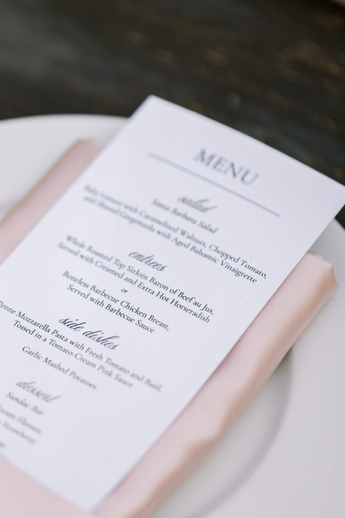 A close-up of a printed menu card placed on a light pink napkin and white plate, featuring sections for salad, entrées, and side dishes in elegant black text. Copyright - Lisa Leanne Photography, California