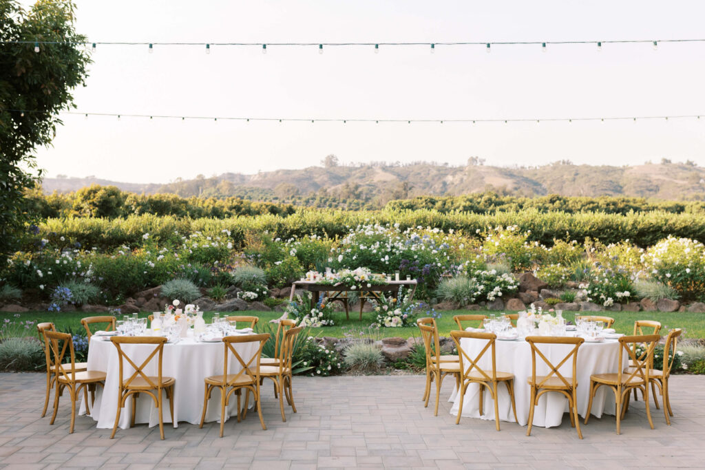 Outdoor event setup with round tables covered in white cloth, wooden chairs, string lights overhead, and a scenic background of green hills, flowering bushes, and trees under a clear sky. Copyright - Lisa Leanne Photography, California