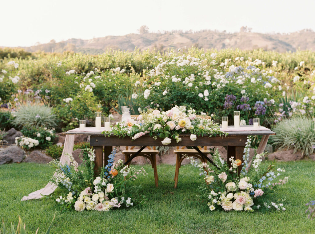 A rustic wooden table set outdoors is decorated with white and pastel flowers, candles, and greenery. Floral arrangements surround the table, with a garden and hills visible in the background. Copyright - Lisa Leanne Photography, California