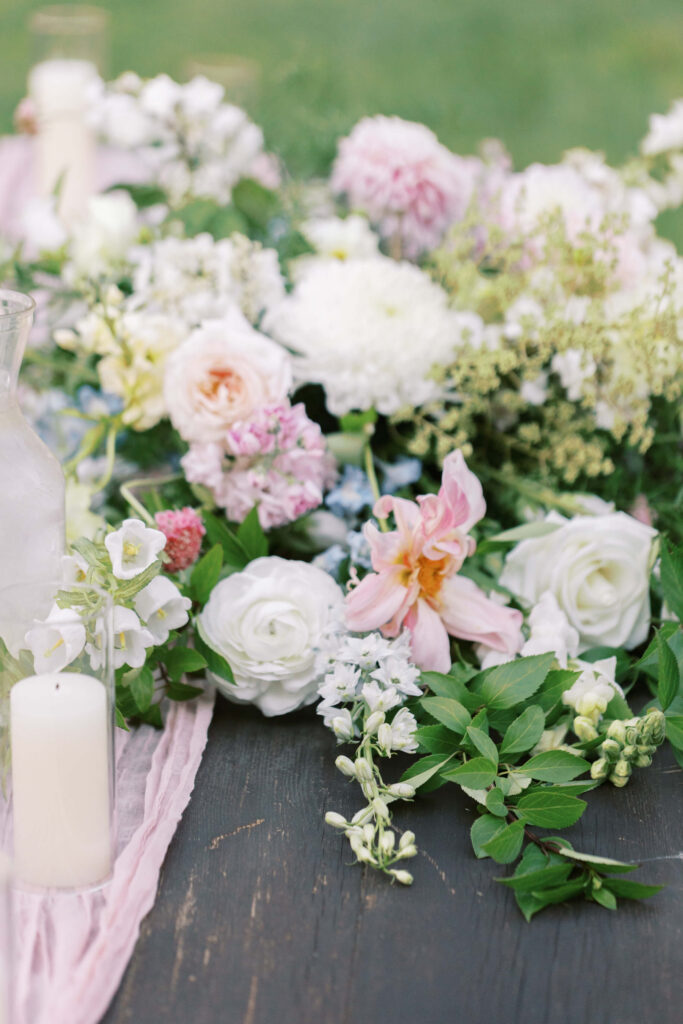 A close-up of a floral arrangement with white, pink, and light purple flowers, green foliage, a white candle, and a glass pitcher on a dark wooden table. The background is blurred. Copyright - Lisa Leanne Photography, California