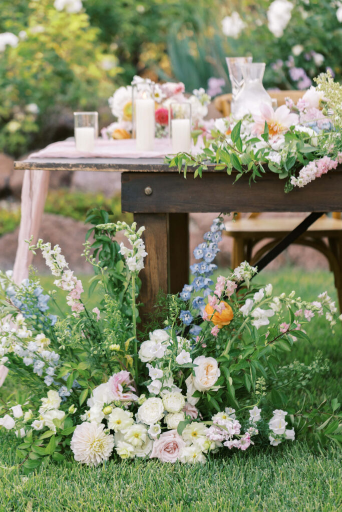A rustic wooden table outdoors is decorated with candles, a glass pitcher, and blush pink, white, and green floral arrangements, with more flowers spilling onto the grass below. Copyright - Lisa Leanne Photography, California