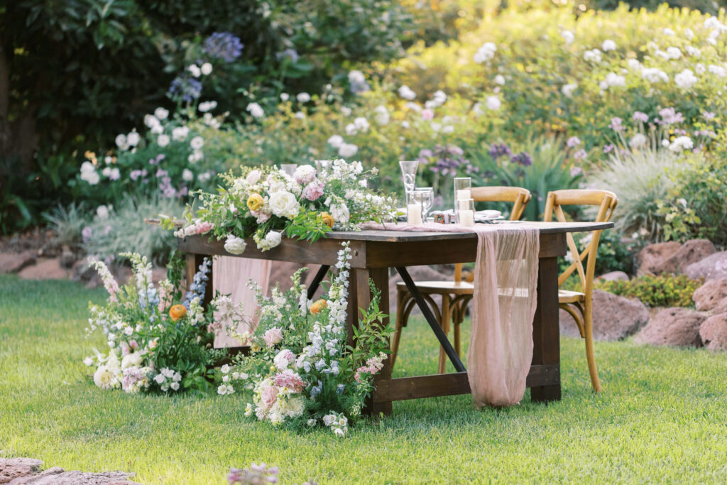 A wooden table decorated with lush flower arrangements and a light pink gauzy table runner stands on green grass, with two wooden chairs in a garden filled with blooming flowers and greenery. Copyright - Lisa Leanne Photography, California
