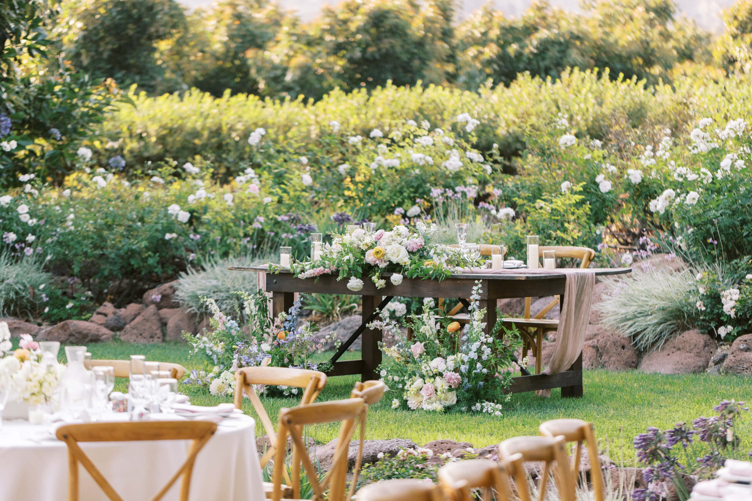 A rustic wooden table decorated with lush floral arrangements and elegant tableware is set outdoors in a garden surrounded by greenery and blooming flowers, with wooden chairs around it. Copyright - Lisa Leanne Photography, California