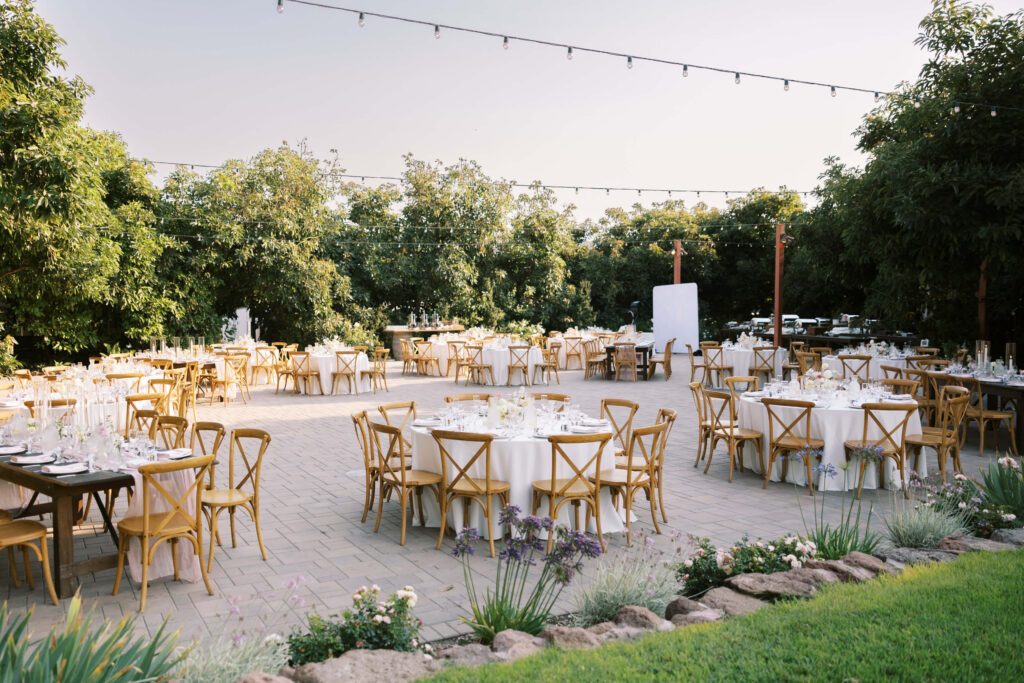 An outdoor event setup with round tables covered in white cloths and wooden chairs on a stone patio, surrounded by greenery and string lights hanging overhead. Copyright - Lisa Leanne Photography, California
