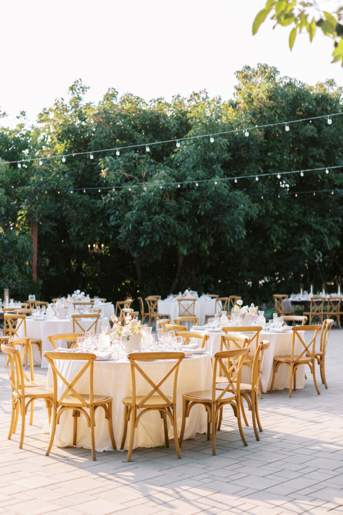 Outdoor event setting with round tables covered in white linens, wooden chairs, floral centerpieces, glassware, and string lights overhead. Lush green trees form a natural backdrop in the background. Copyright - Lisa Leanne Photography, California