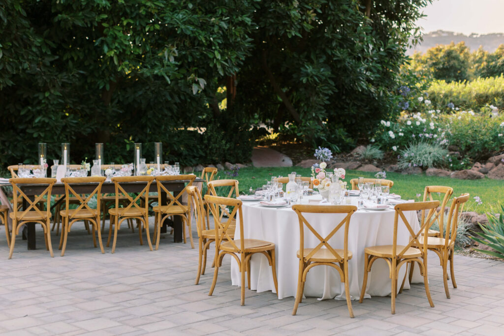 An outdoor dining setup with wooden chairs and tables covered in white tablecloths, set for a meal with plates, glasses, and floral centerpieces, surrounded by greenery and trees. Copyright - Lisa Leanne Photography, California