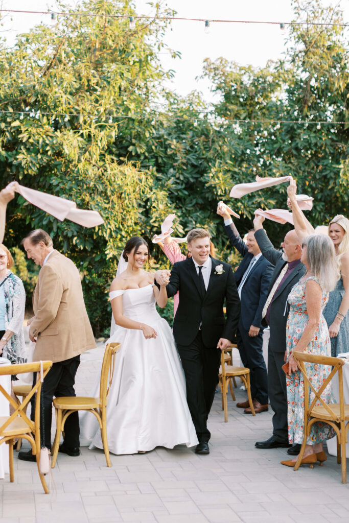 A bride and groom walk hand in hand down an outdoor aisle, smiling as guests on both sides wave napkins in celebration. The scene is festive with trees in the background and chairs along the aisle. Copyright - Lisa Leanne Photography, California