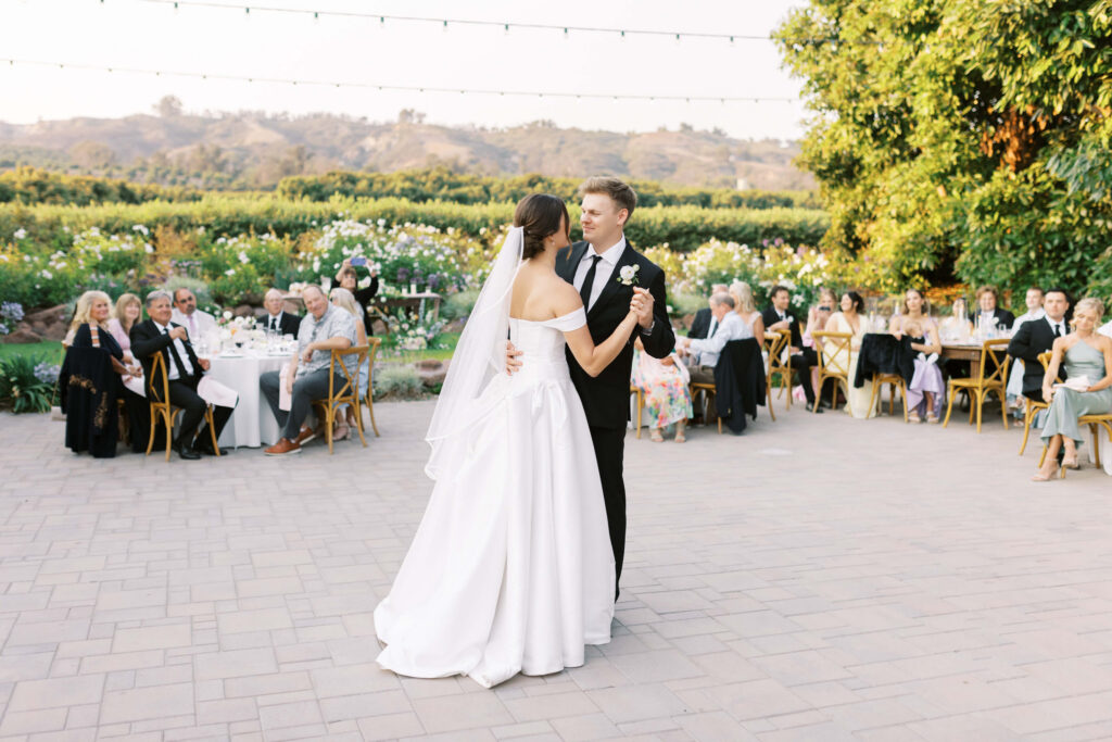 A bride and groom share their first dance outdoors on a patio, surrounded by seated guests at tables, with greenery and string lights in the background on a sunny day. Copyright - Lisa Leanne Photography, California