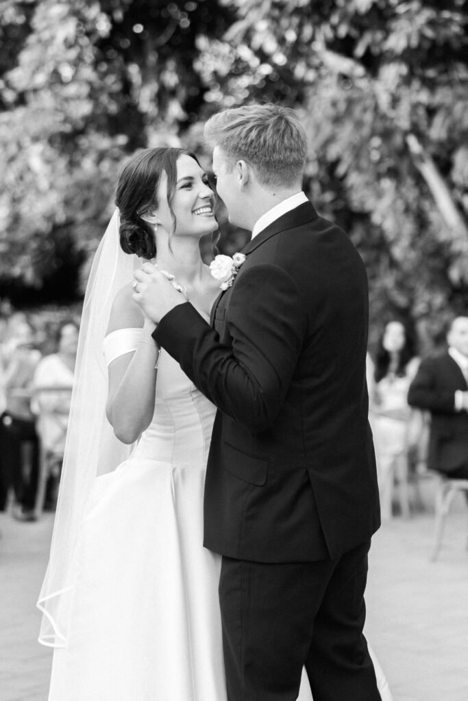 A bride and groom share a joyful first dance outdoors. The bride wears an off-shoulder wedding gown and veil, while the groom is in a dark suit. They smile at each other, surrounded by seated guests and leafy trees. Copyright - Lisa Leanne Photography, California