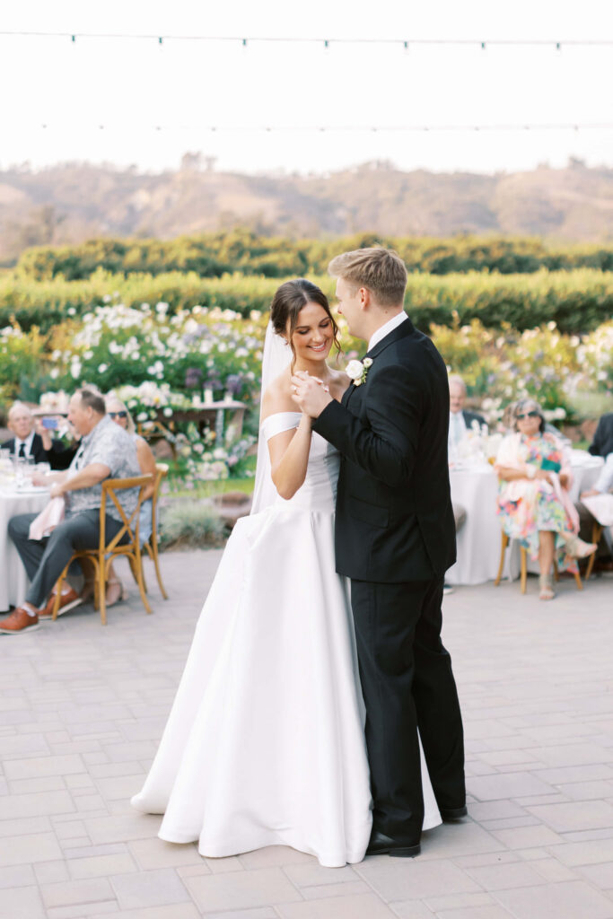 A bride and groom share their first dance outdoors at their wedding reception, surrounded by guests seated at tables. Lush greenery and hills are visible in the background under string lights. Copyright - Lisa Leanne Photography, California