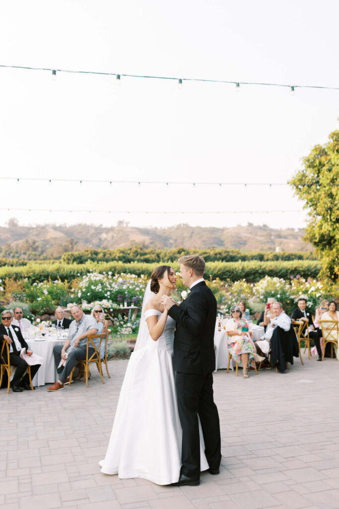 A bride in a white dress and a groom in a black suit share their first dance outdoors, surrounded by seated guests at tables, with greenery and string lights in the background. Copyright - Lisa Leanne Photography, California