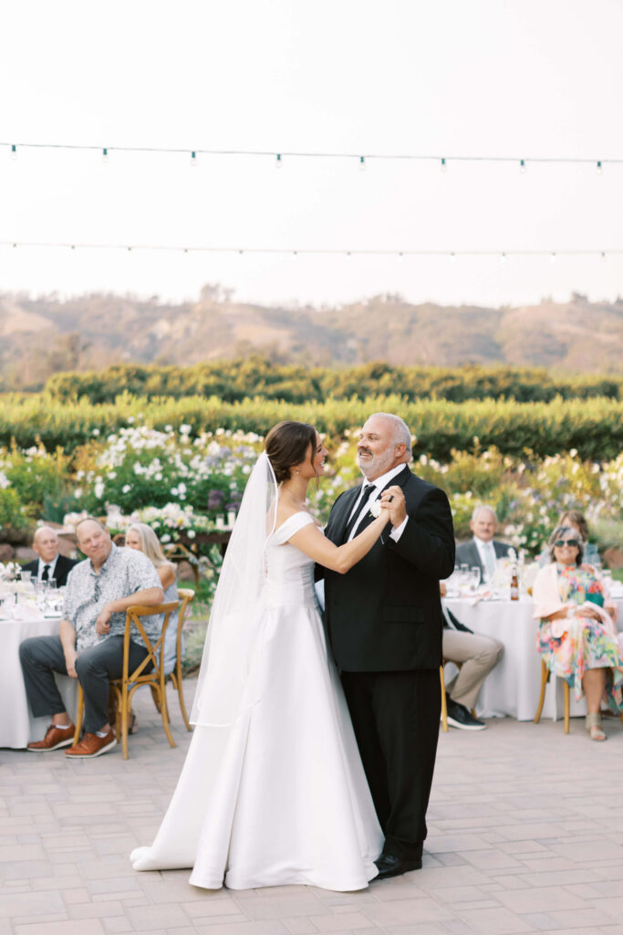 A bride in a white gown and veil dances with an older man in a black suit outdoors, surrounded by seated guests at round tables with greenery and hills in the background. Copyright - Lisa Leanne Photography, California
